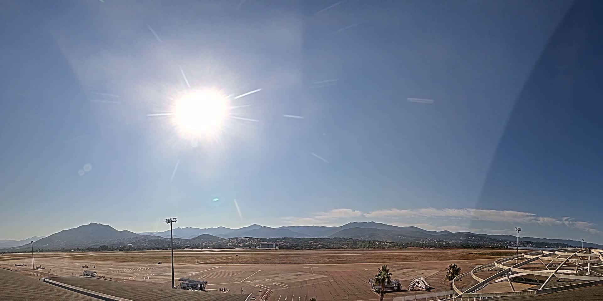 A sunny day reveals a tarmac with airport equipment, extending to distant mountains under a mostly clear blue sky.