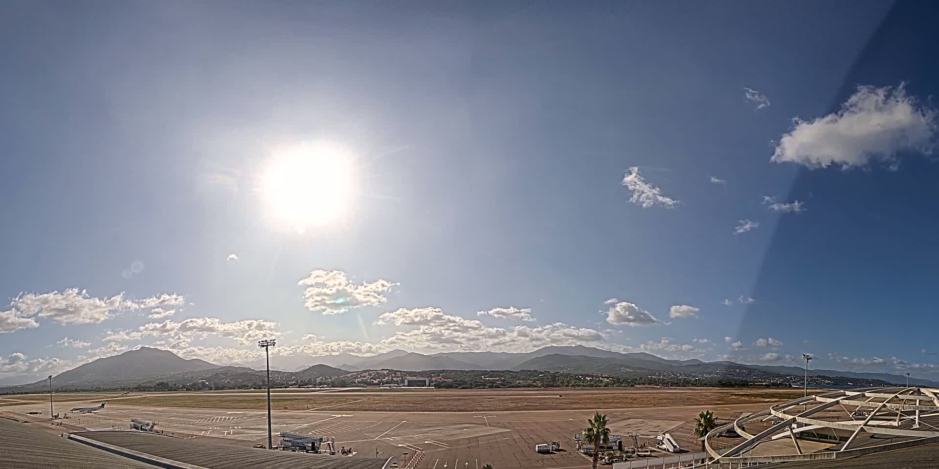 A sunny day with scattered clouds shows an airport tarmac, runway, and distant mountains.