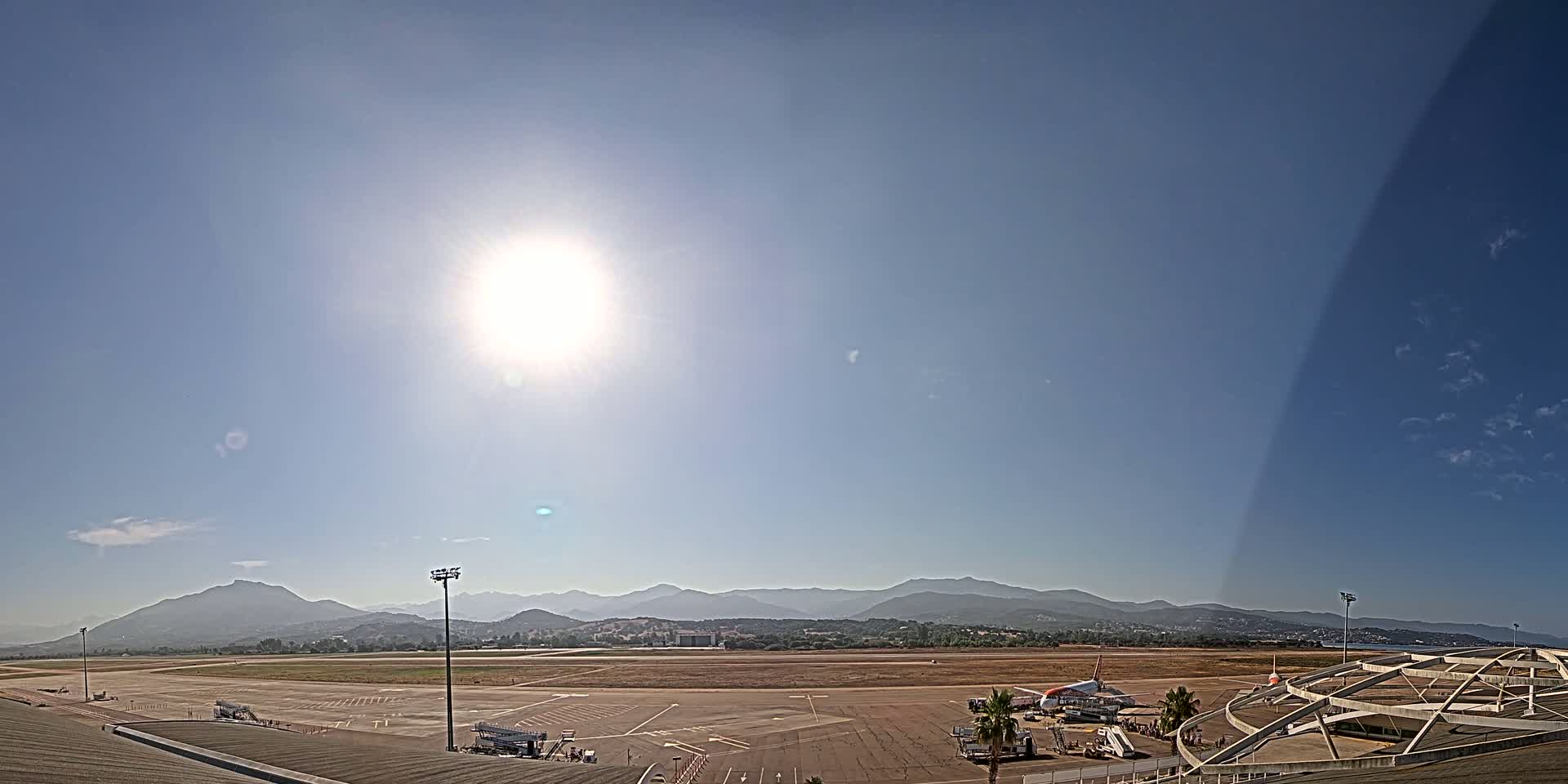 A sunny day at an airport shows an airplane on the tarmac with mountains in the background.