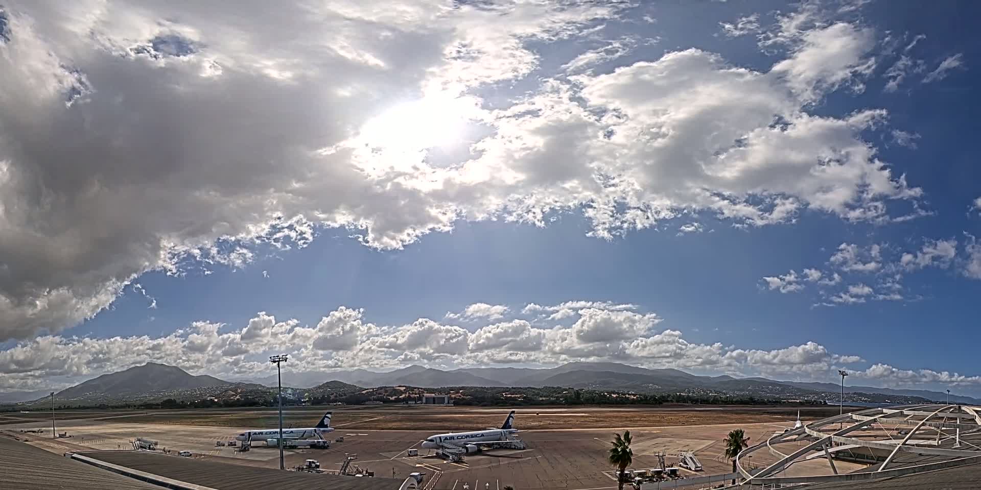 Two airplanes are parked on an airport tarmac under a partly cloudy sky with mountains in the background.