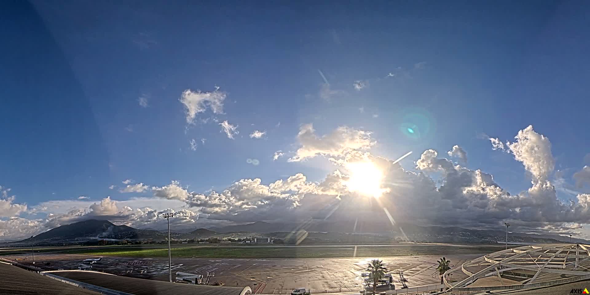 A bright sun shines through scattered cumulus clouds over a wet airport tarmac with distant mountains, suggesting clear weather after recent rain.