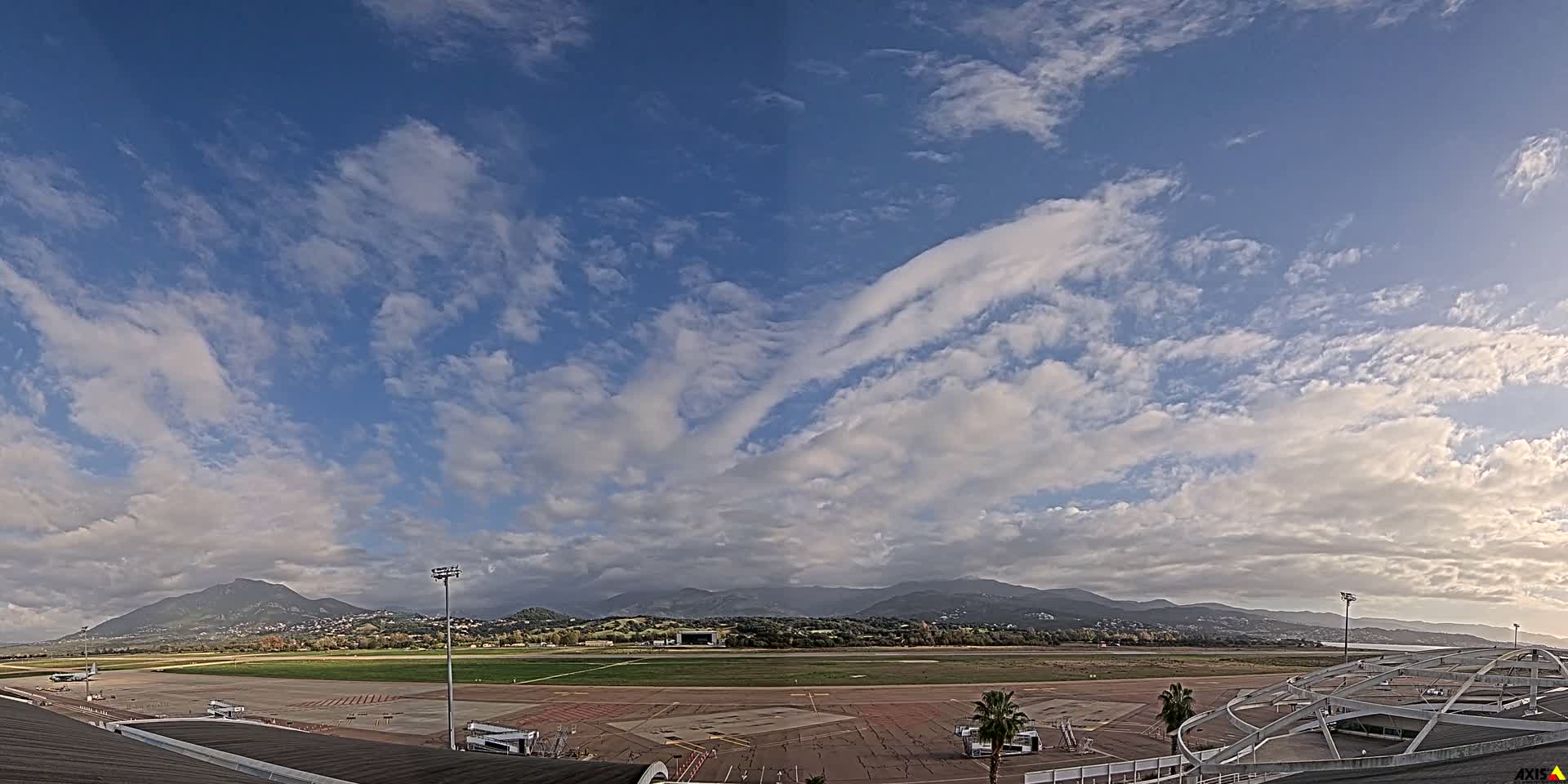 A bright sun shines through scattered cumulus clouds over a wet airport tarmac with distant mountains, suggesting clear weather after recent rain.