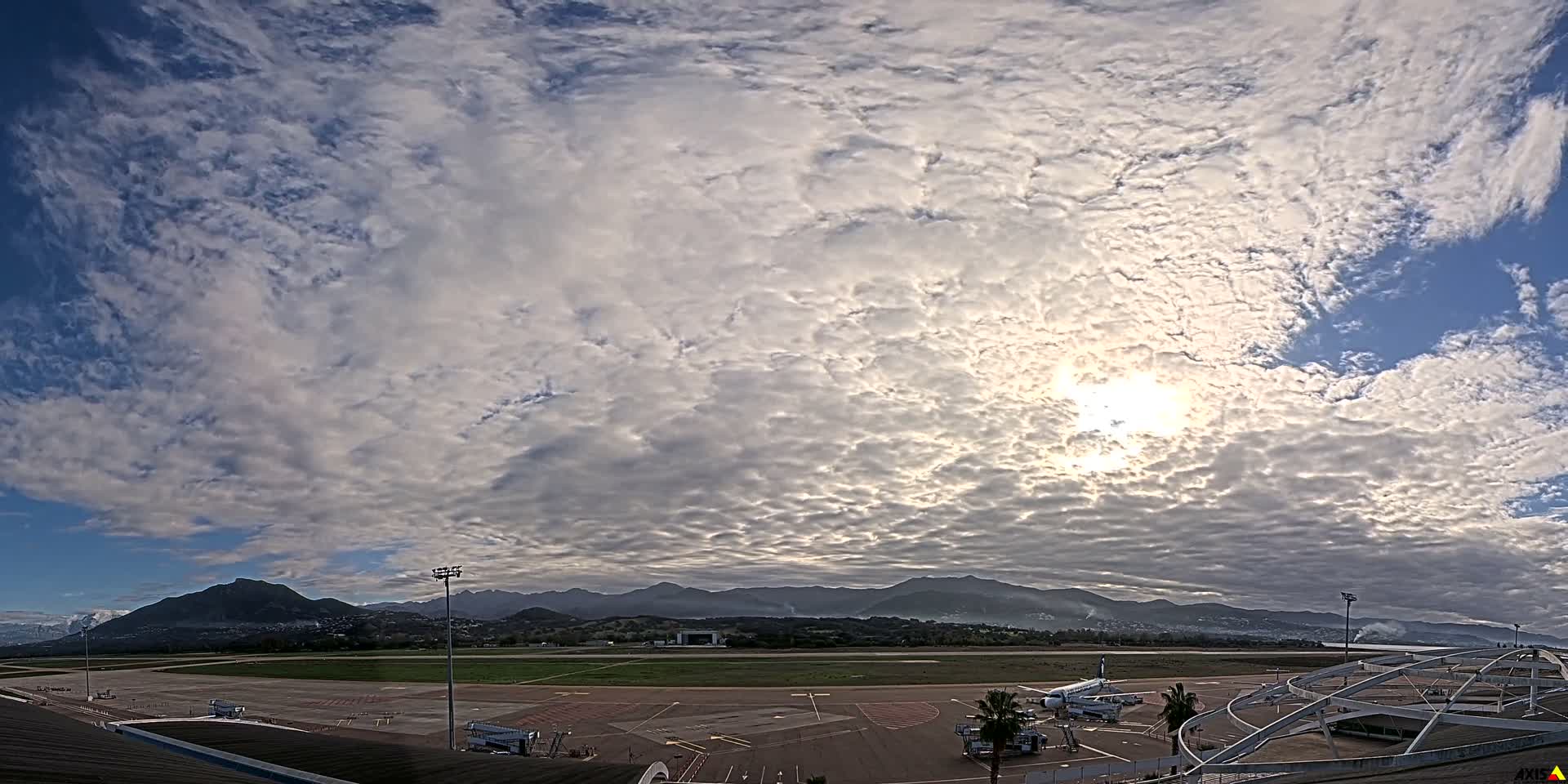 An expansive view captures an airport tarmac with parked airplanes and distant mountains under a partly cloudy sky with bright sunshine.