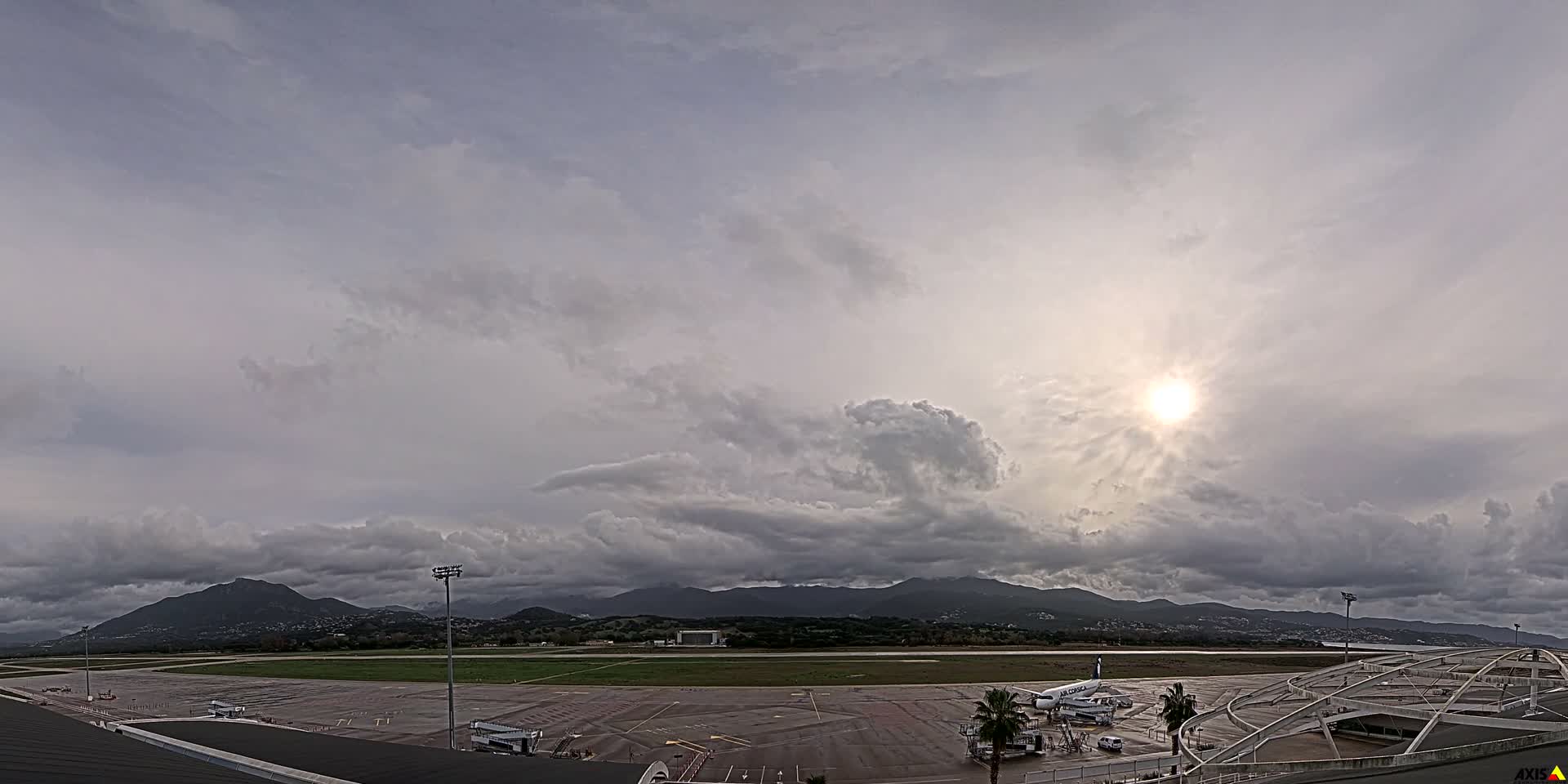 An expansive view of an airport tarmac with parked airplanes and a runway stretches towards distant mountains and a body of water, all beneath a heavily clouded sky with the sun brightly peeking through on the right.