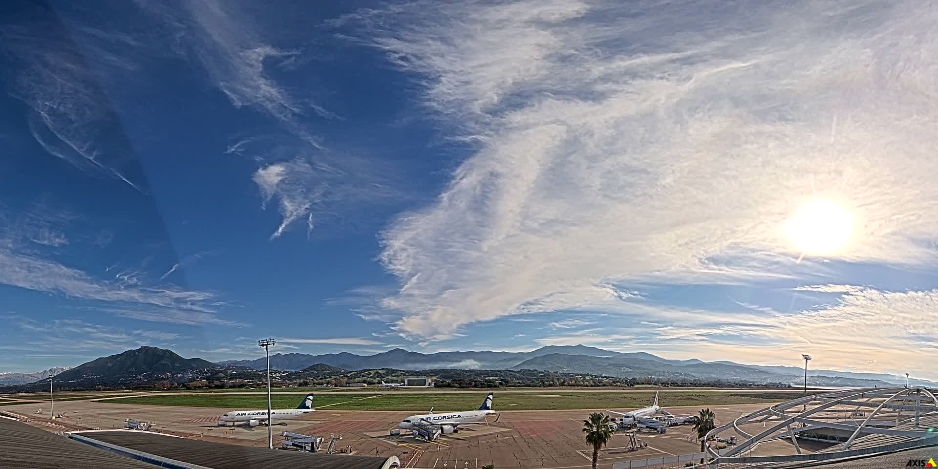 An expansive view of an airport tarmac with parked airplanes and a runway stretches towards distant mountains and a body of water, all beneath a heavily clouded sky with the sun brightly peeking through on the right.