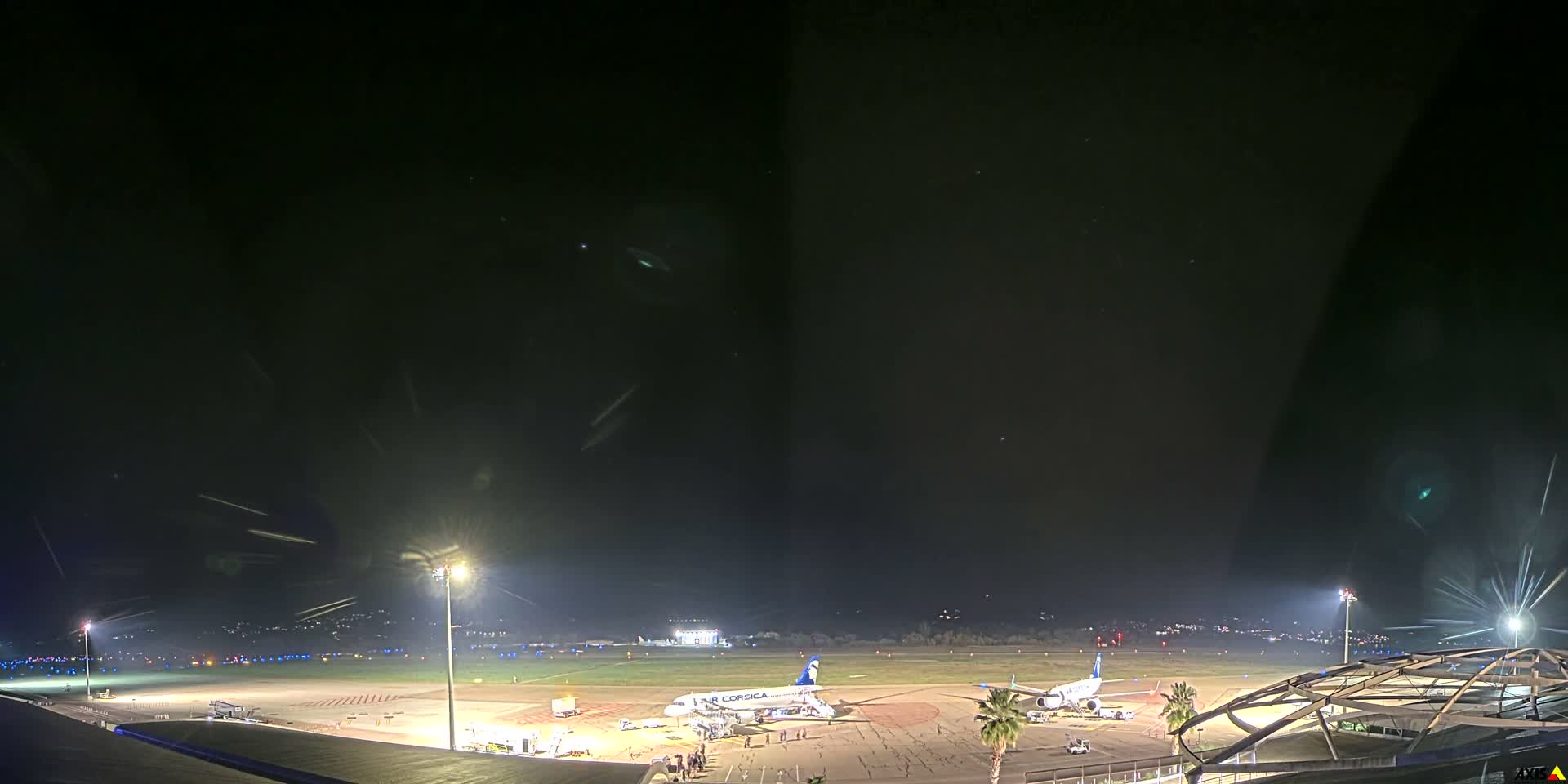 An expansive view of an airport tarmac with parked airplanes and a runway stretches towards distant mountains and a body of water, all beneath a heavily clouded sky with the sun brightly peeking through on the right.