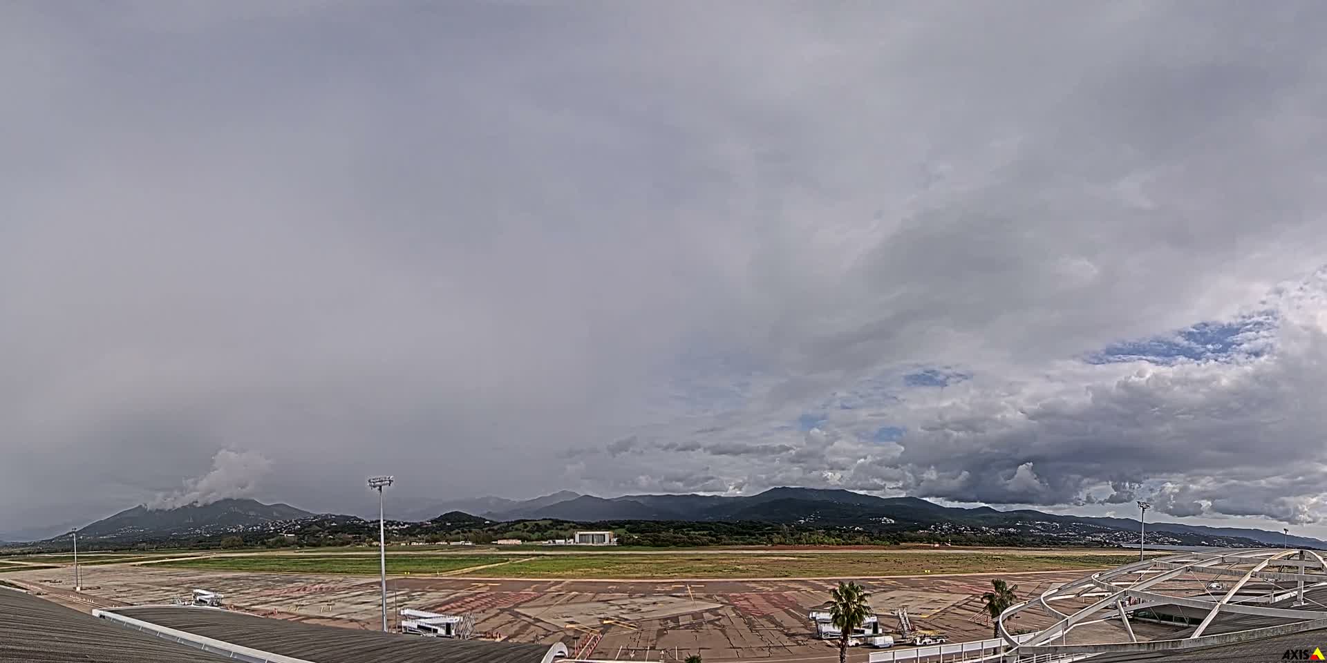 An expansive view of an airport tarmac with parked airplanes and a runway stretches towards distant mountains and a body of water, all beneath a heavily clouded sky with the sun brightly peeking through on the right.