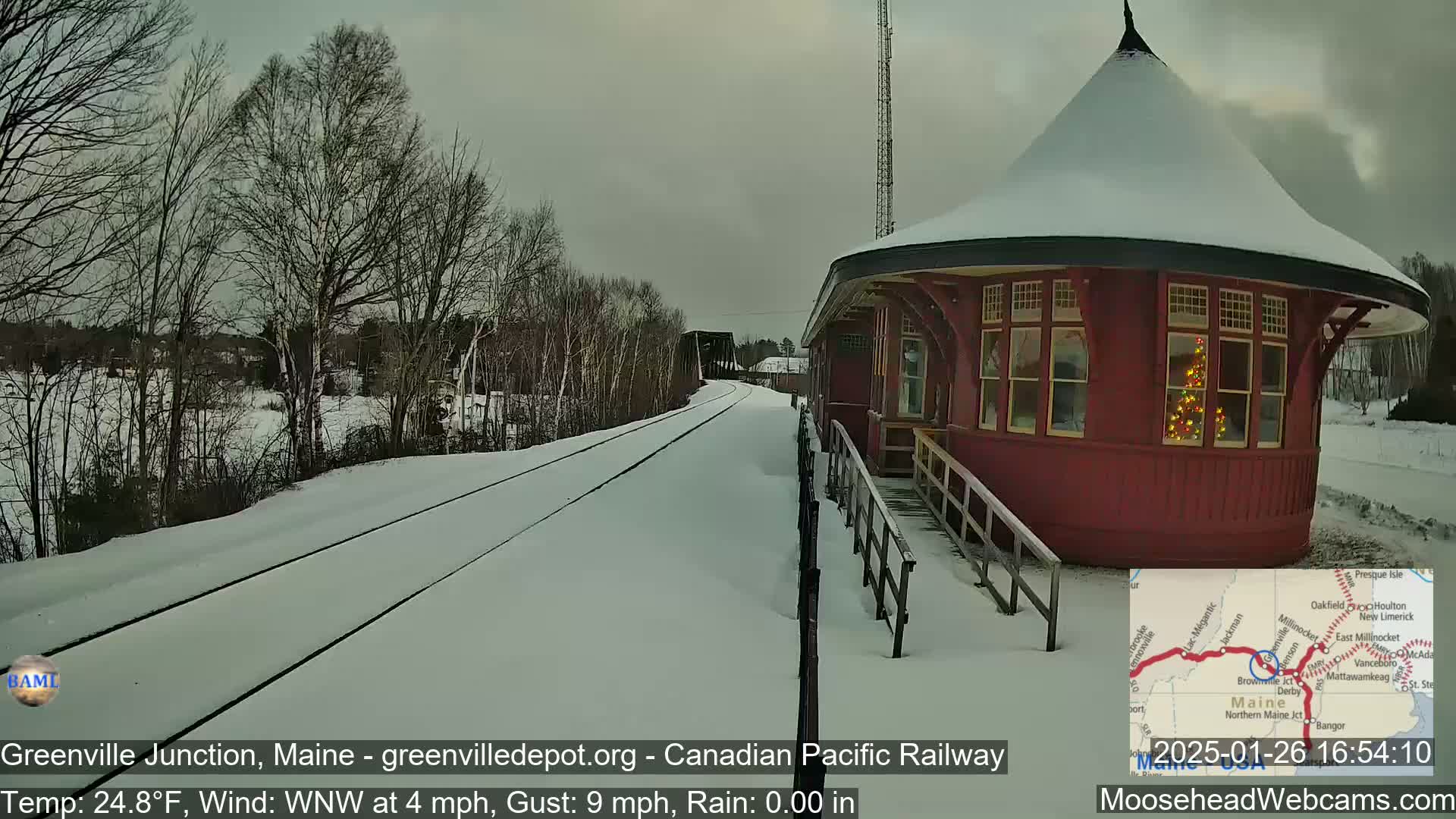 A snow-covered train station, next to a railway track and surrounded by bare winter trees, is seen under an overcast sky.