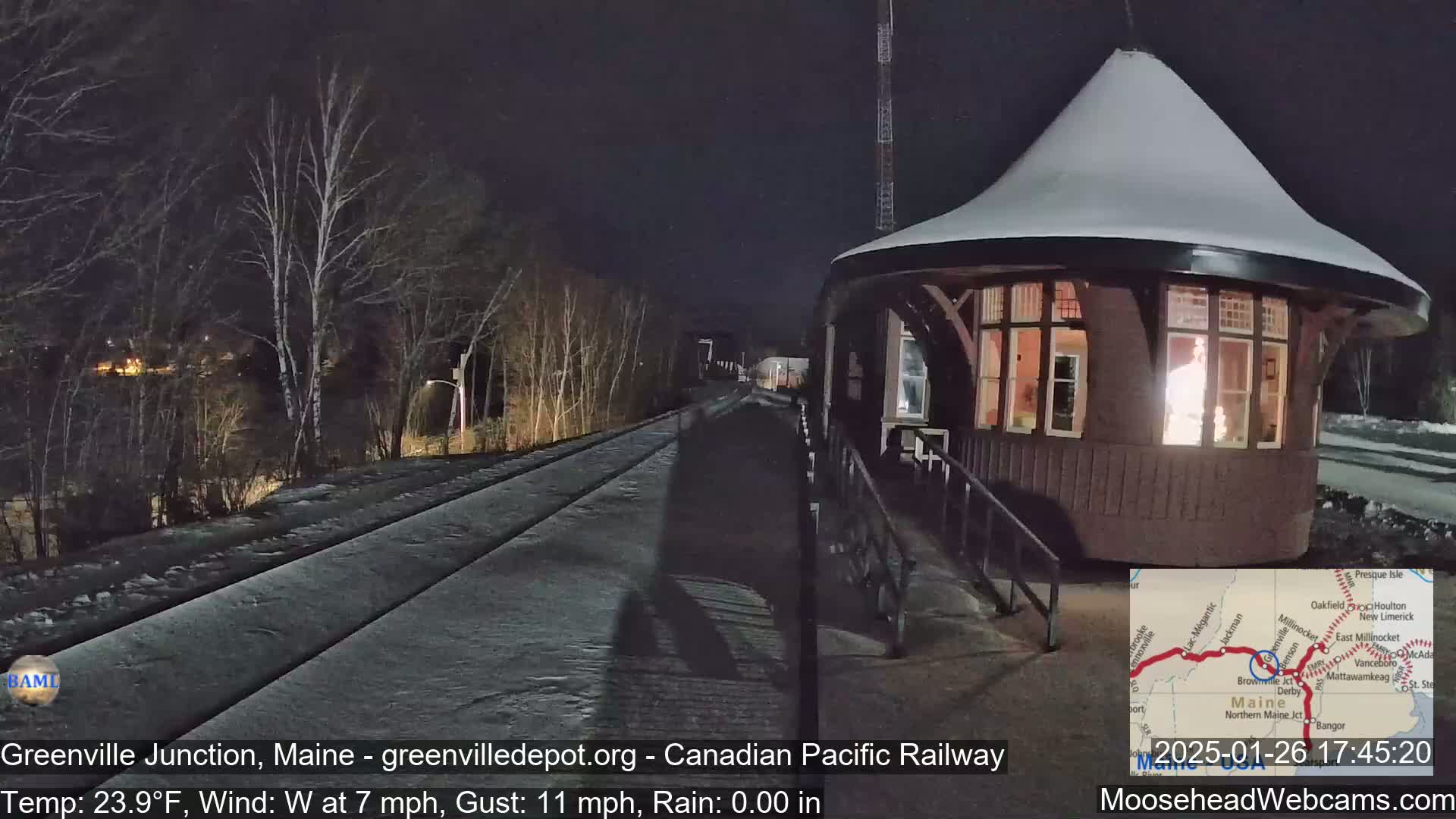 A snow-covered train depot is illuminated at night next to a snow-covered railway track under a clear, dark sky.
