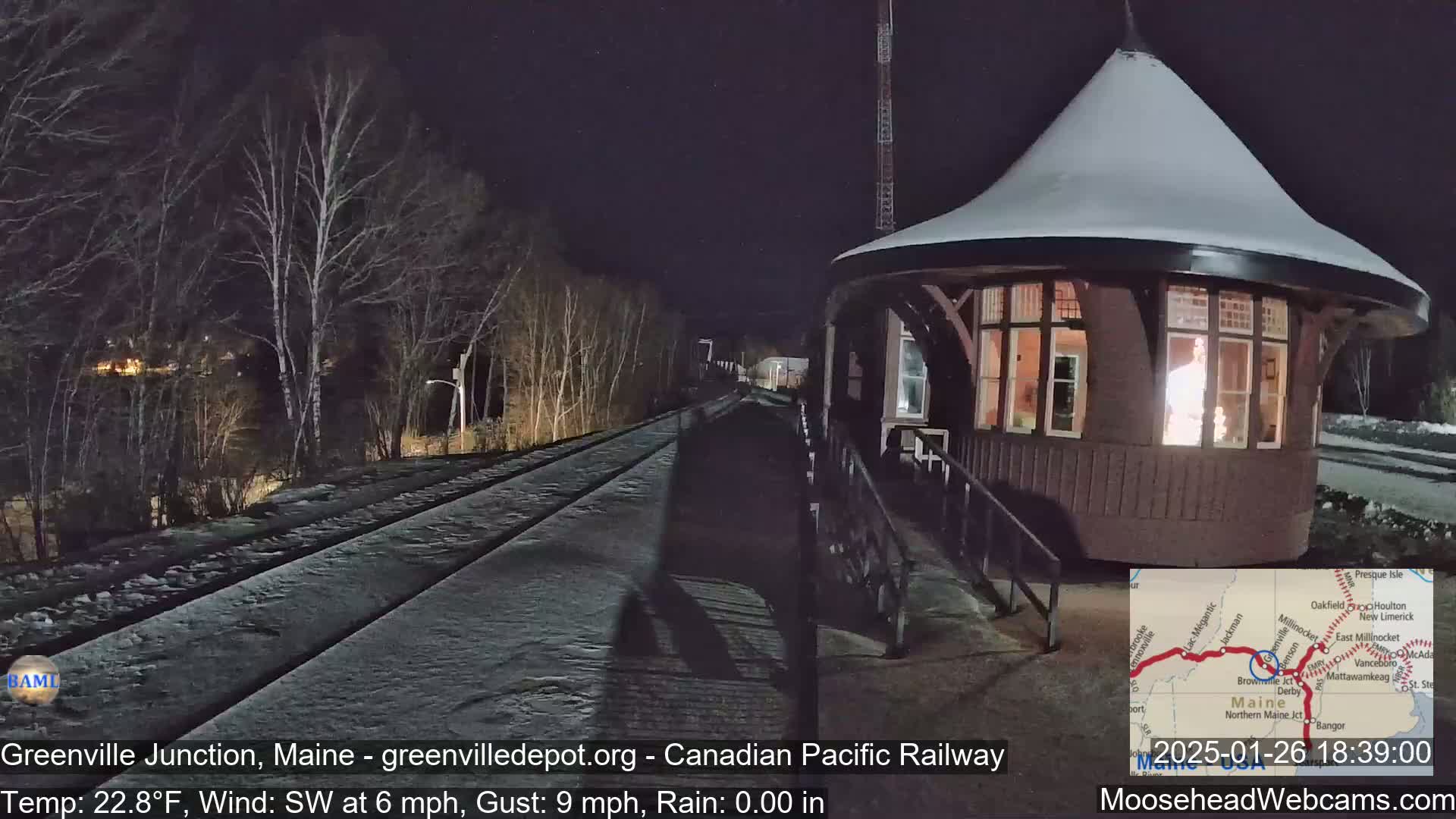A snow-covered train platform with a small, illuminated round building is visible at night, under a dark sky with a southwest wind.