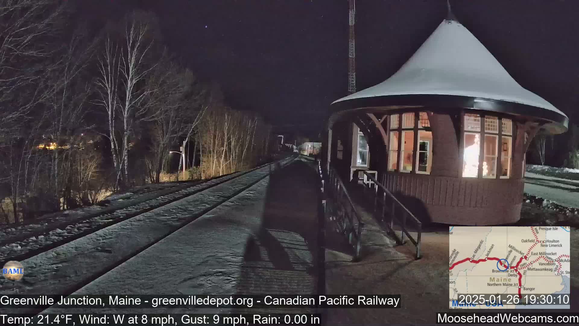 A snow-covered train platform with a lit-up waiting room is seen at night, under a clear sky with a westerly wind.