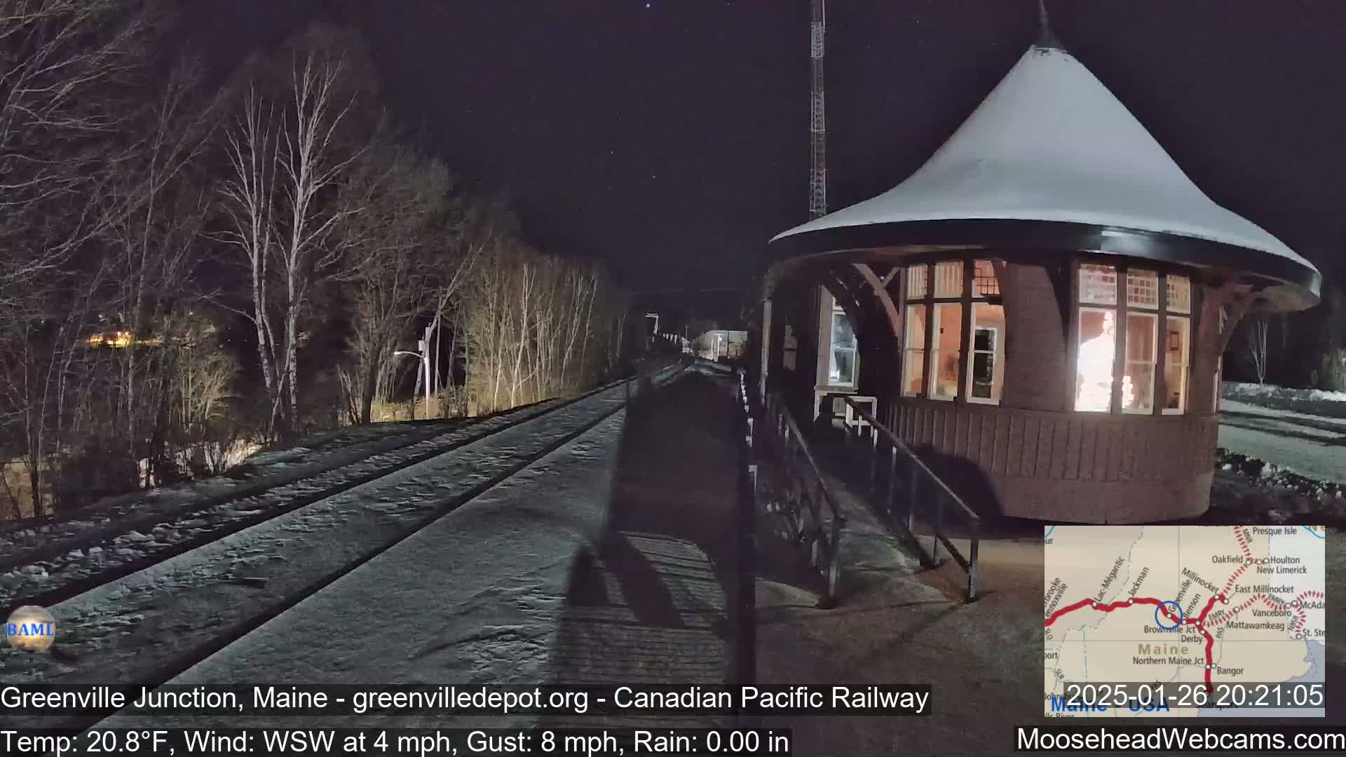 A snow-covered train station with illuminated windows sits beside a snow-covered railway track at night under a dark sky.