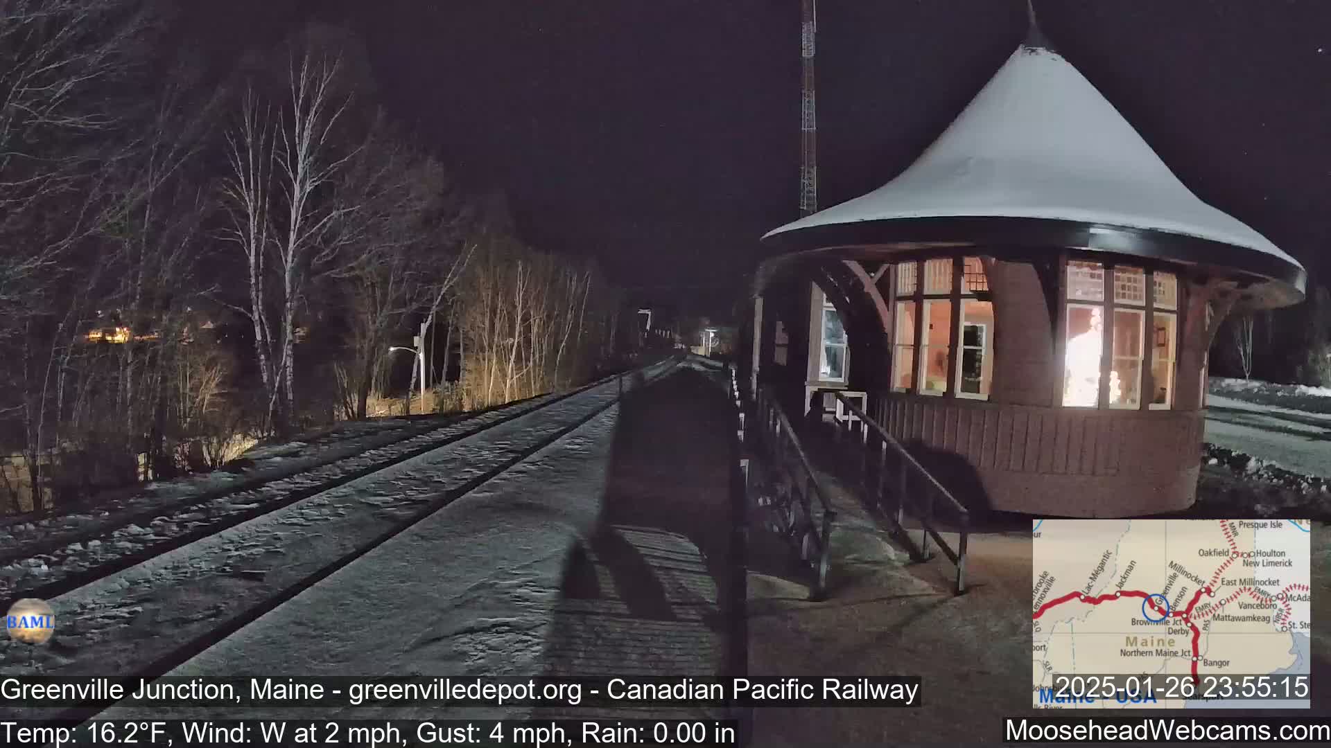 A snow-covered train station, illuminated at night, sits beside a snow-covered railway track under a dark sky.