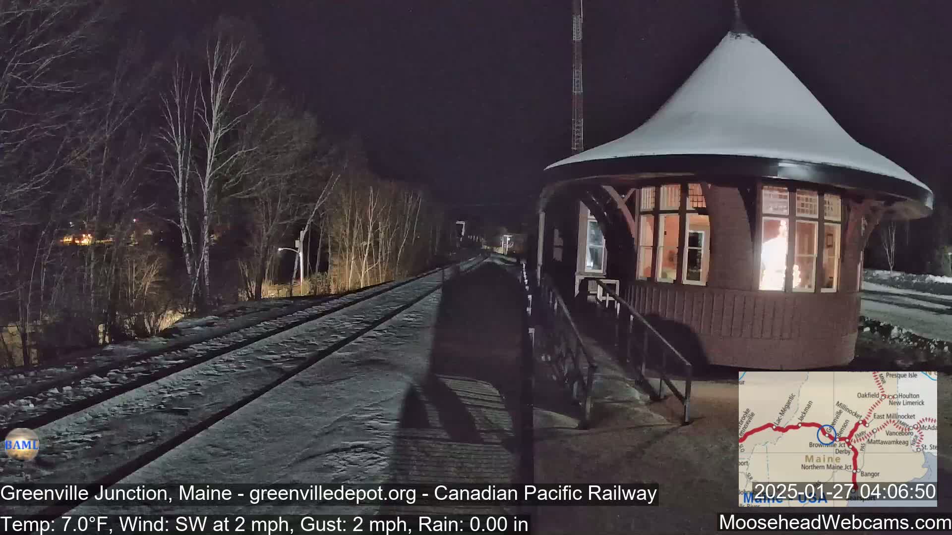 A snow-covered train platform with a round, illuminated depot building is visible at night, under a clear sky with a temperature of 7 degrees Fahrenheit and a light southwest wind.