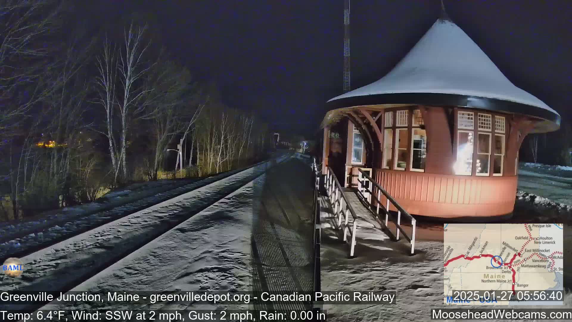 A snow-covered railway station, illuminated at night, is situated beside a snow-covered railway track under a dark, clear sky.