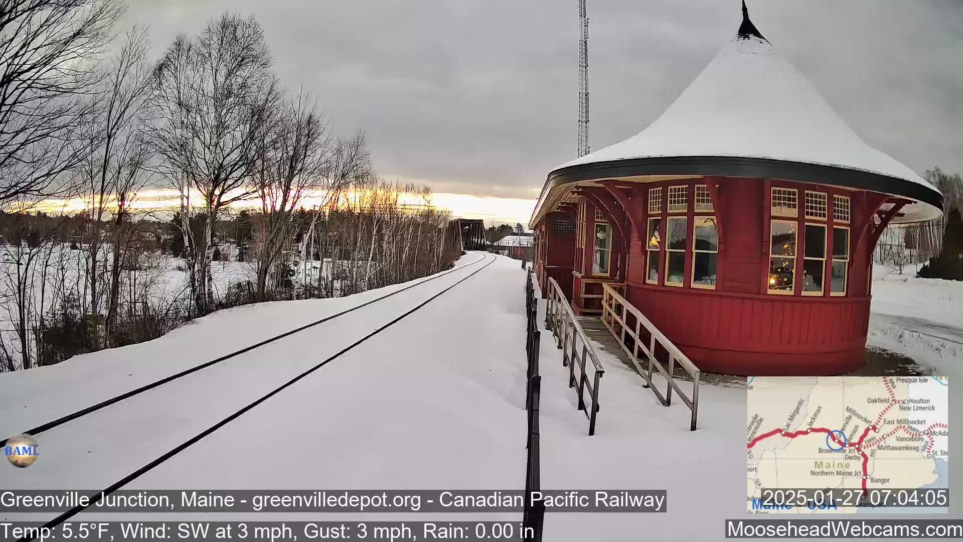 A red octagonal train depot sits beside snow-covered train tracks during a sunset under a mostly cloudy sky.
