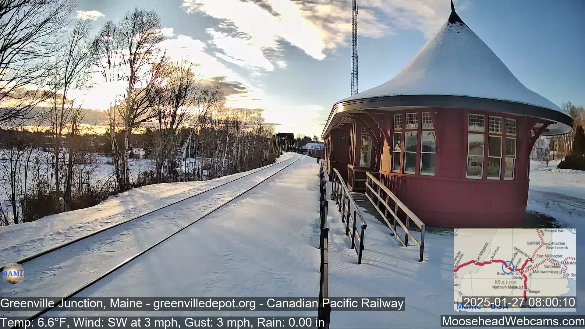 A snow-covered train platform with a red octagonal building is visible at sunrise, with a railway extending into the distance under a partly cloudy sky.