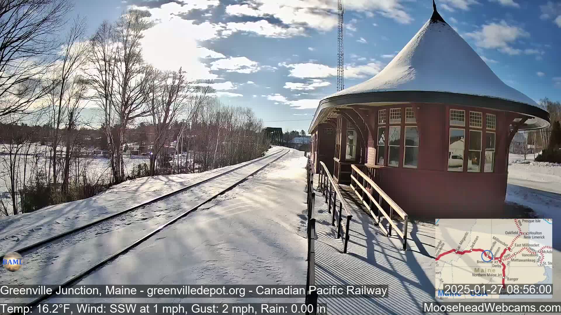 A snow-covered train platform with a red, octagonal train station building is visible under a partly sunny sky, with a light south-southwesterly wind.