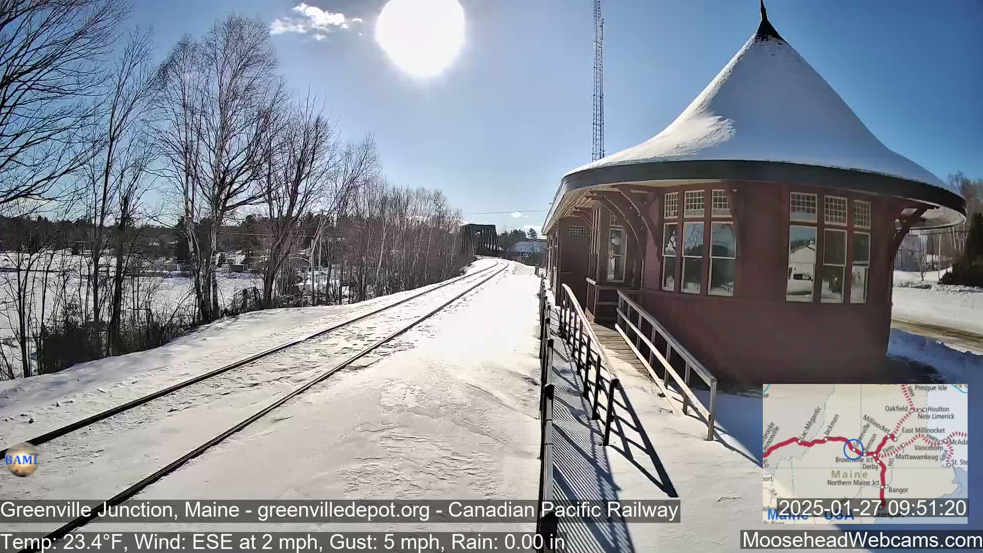A snow-covered train platform with a round, reddish-brown train station building is under a bright, sunny sky.