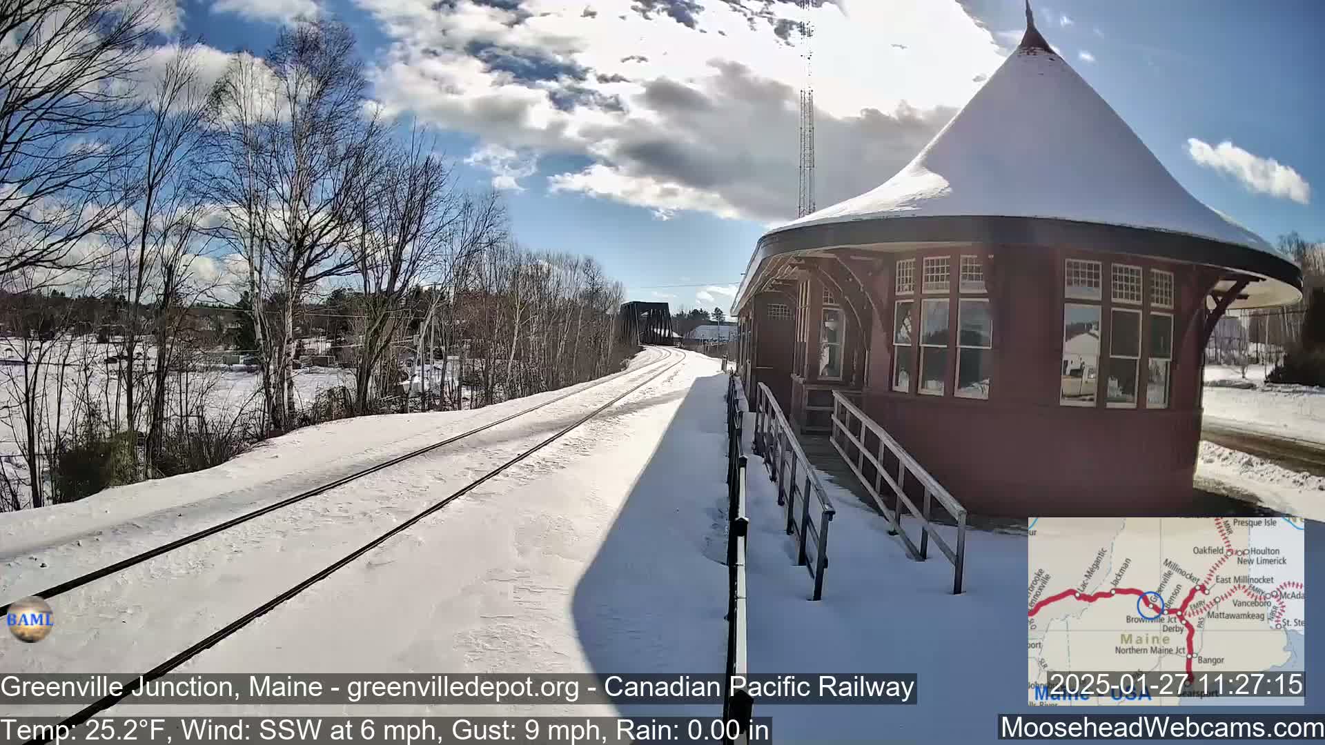 Snow-covered train tracks extend from a snow-dusted, round train depot under a partly sunny sky.
