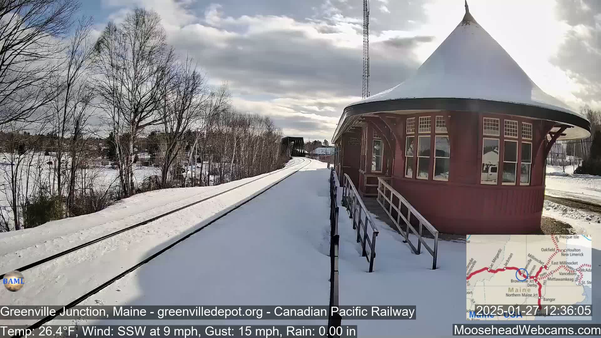 A snow-covered train track leads to a red, octagonal train depot under a partly cloudy sky.