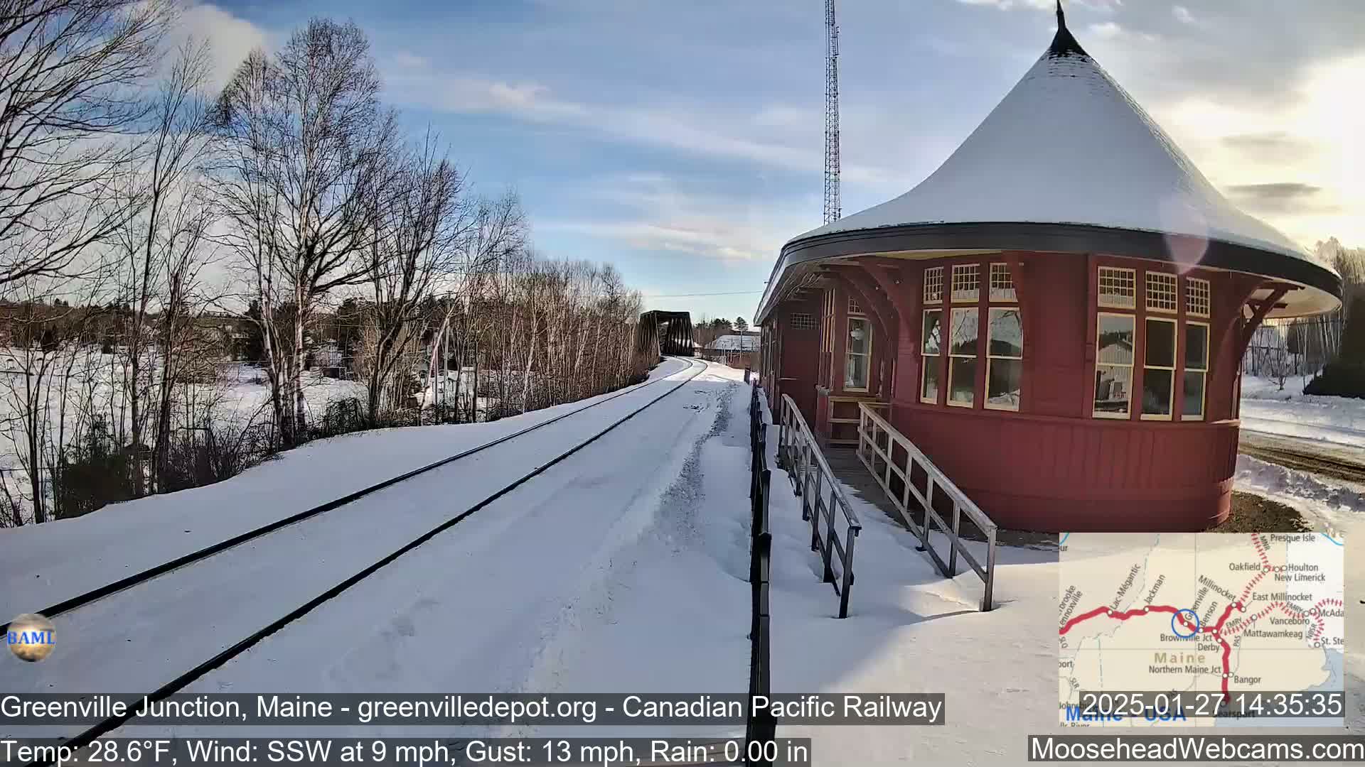 A snow-covered train station with a round, red building and nearby railroad tracks under a partly sunny sky.