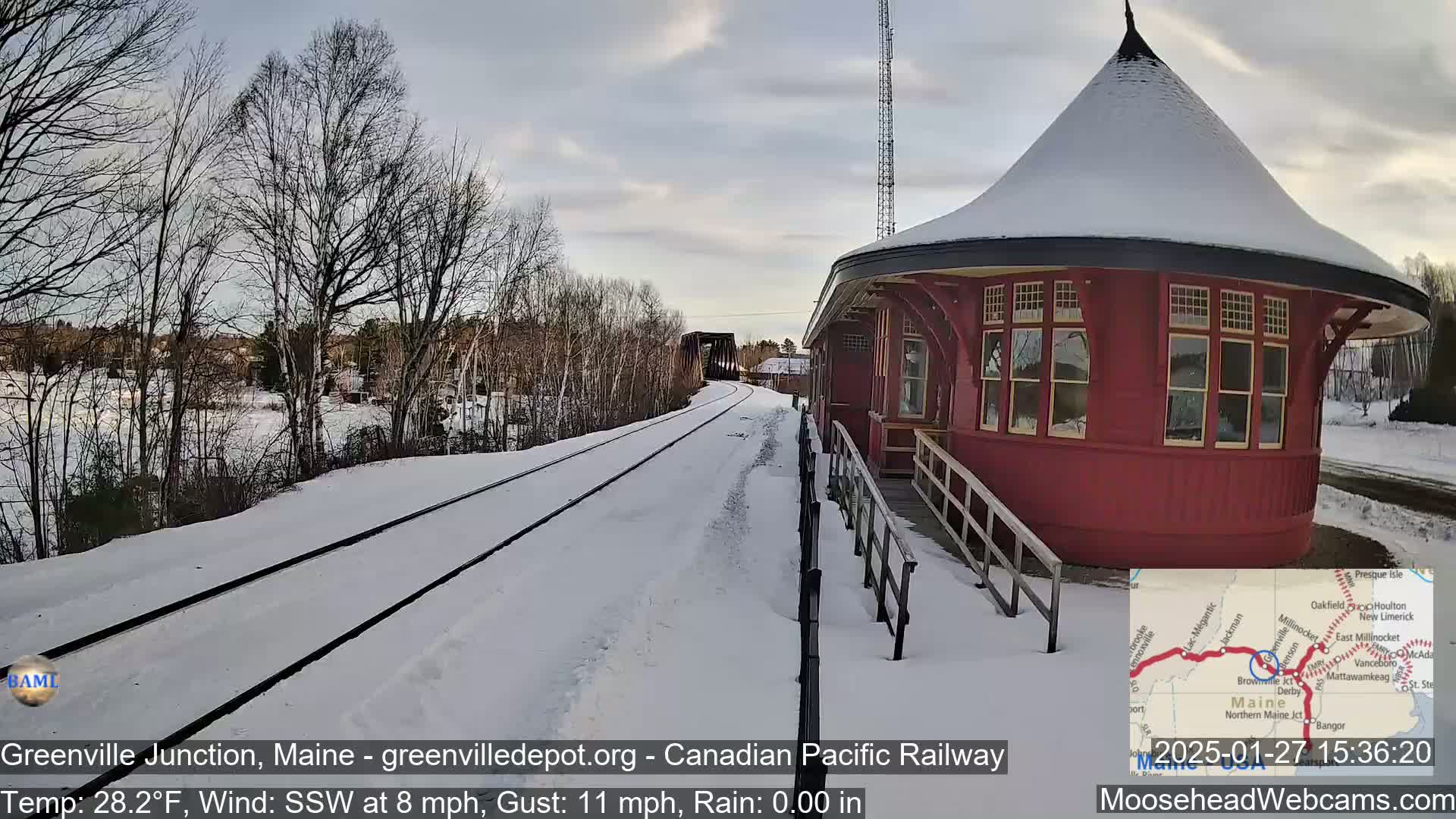 A snow-covered train track leads to a red, round train depot next to bare winter trees under a cloudy sky.