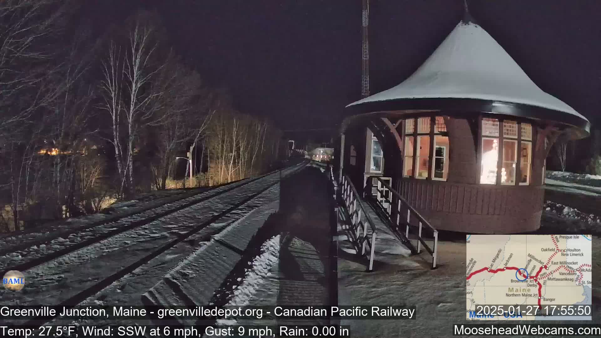 A snow-covered train platform with a round, illuminated station building is seen at night under a dark sky, with a south-southwest wind at 6 mph and gusts up to 9 mph.
