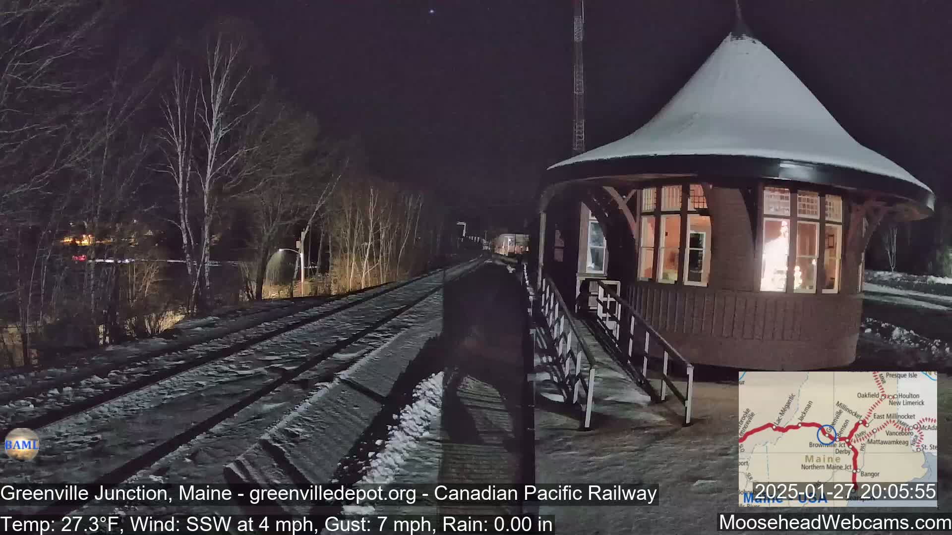 A snow-covered train station platform and railway tracks are visible at night under a clear sky, with a small building illuminated in the foreground.