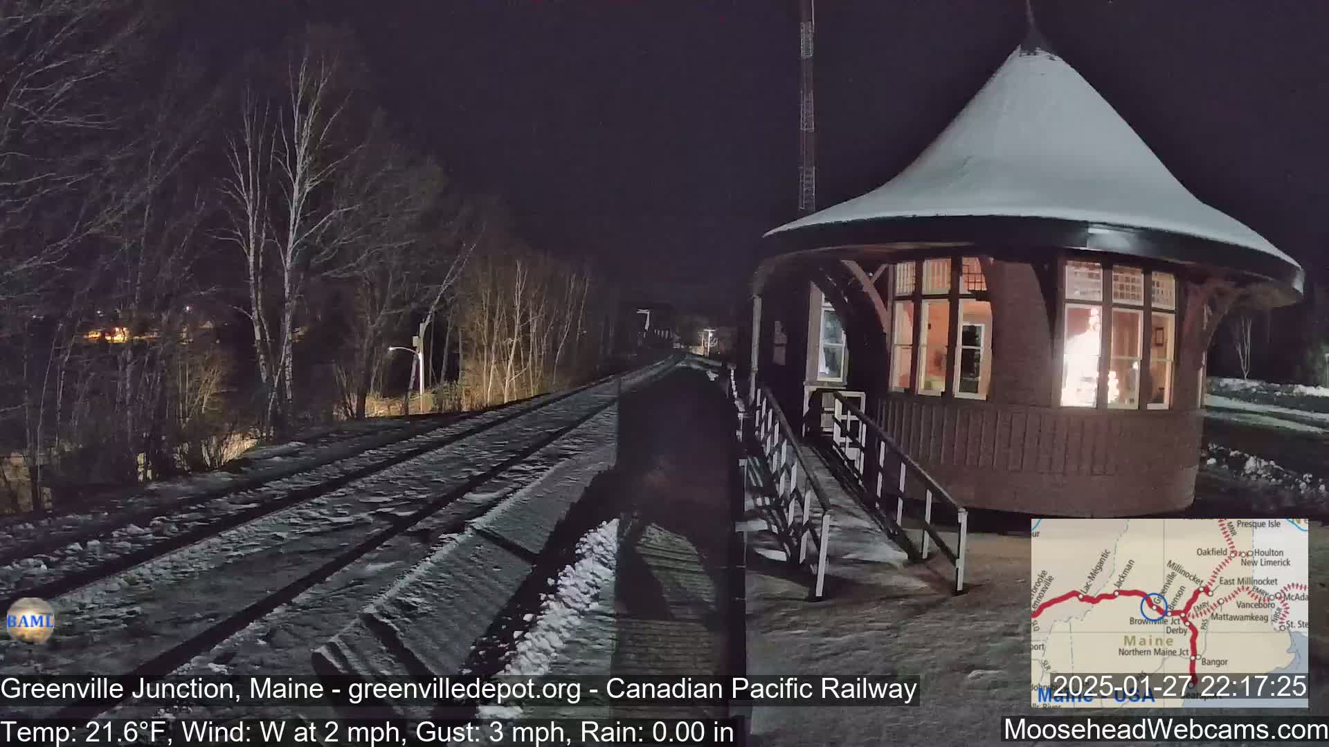 A snow-covered train platform with a round, illuminated building is seen at night, under a clear sky with a westerly wind.