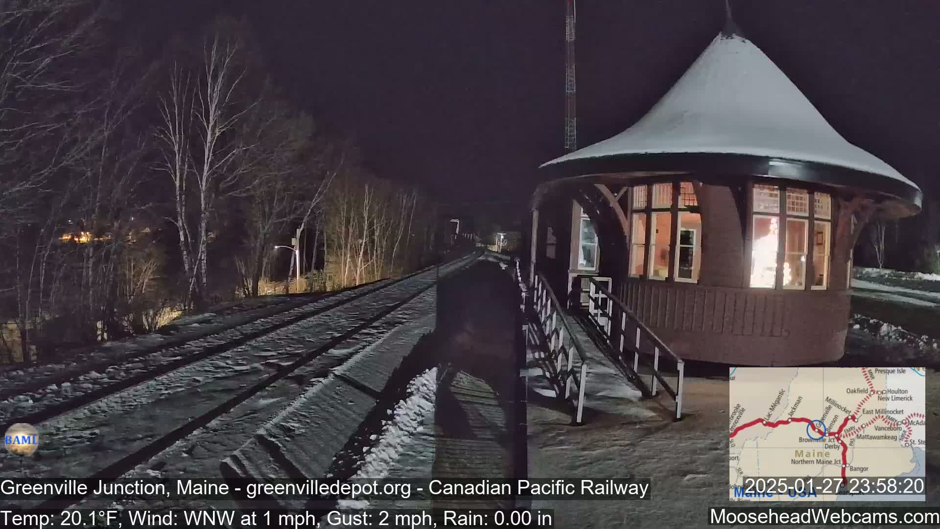 A snow-covered, illuminated train depot stands beside a snow-covered railway track at night, under a dark sky with a west-northwest wind.