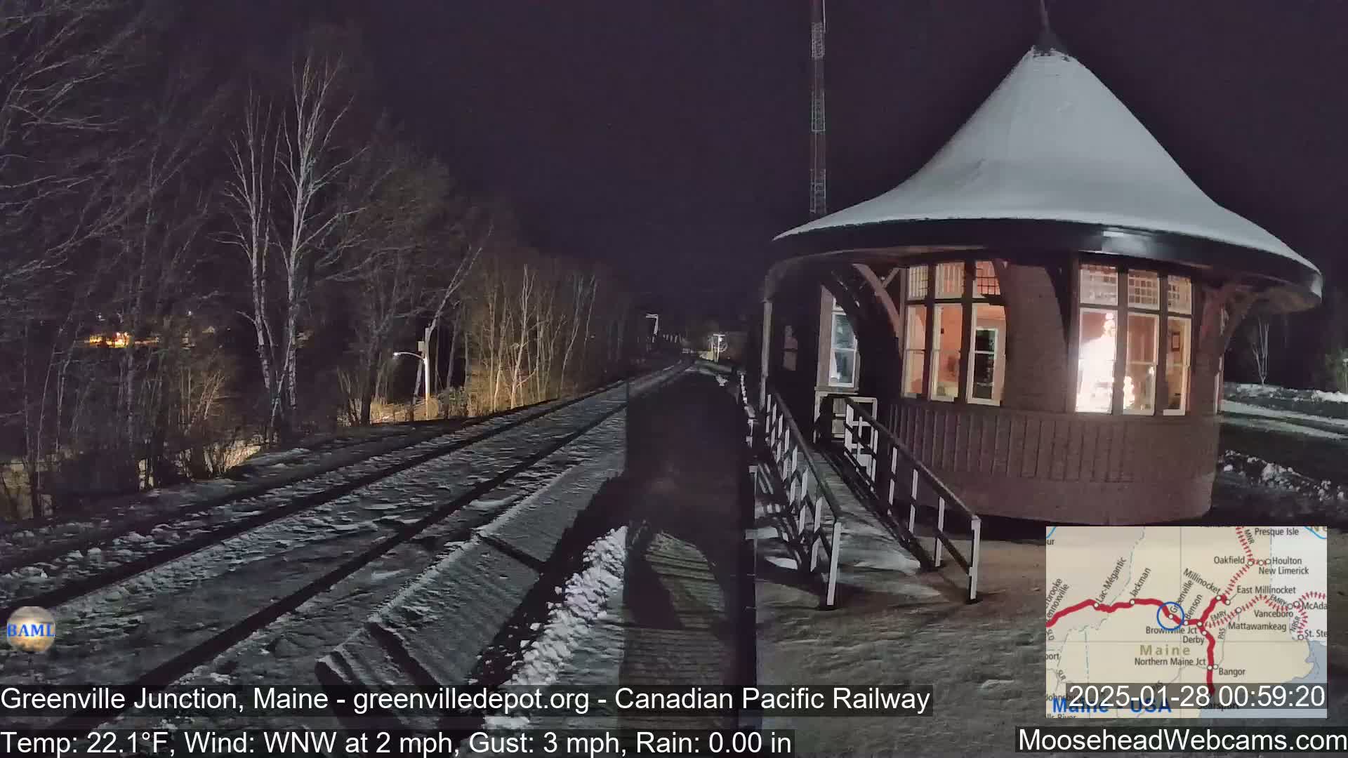 A snow-covered railway track leads to a small, illuminated, snow-covered building at night.