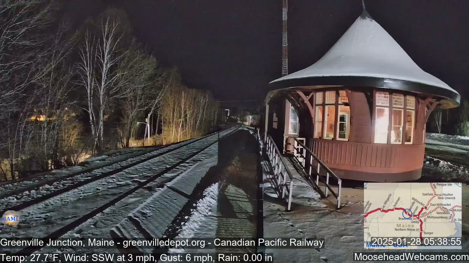 A snow-covered train station, illuminated at night, sits beside a snow-covered railway track that leads into a dark, snow-covered wooded area under a dark night sky.