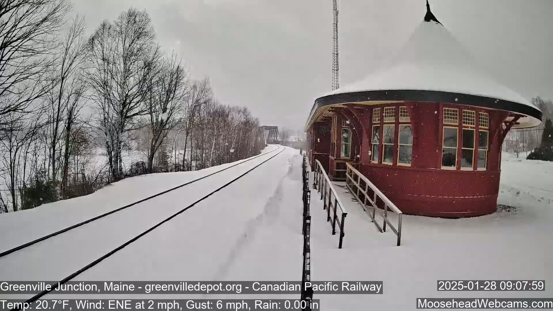 Snow falls on a snow-covered railroad track next to a round, red train station.