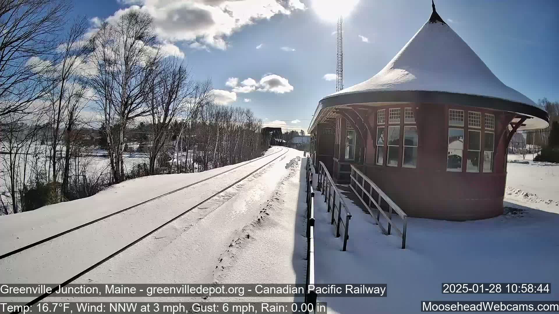 Snow-covered train tracks lead to a snow-dusted, round train station under a sunny sky with some clouds.