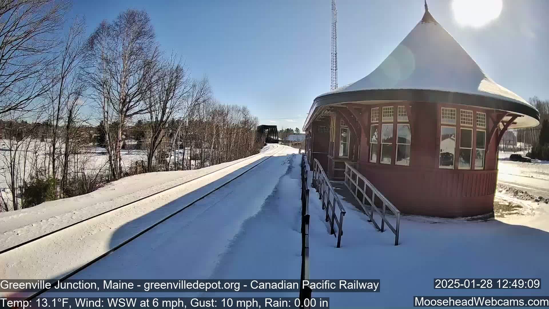 A snow-covered train platform with a red, round train station and train tracks extends into the distance under a sunny winter sky.