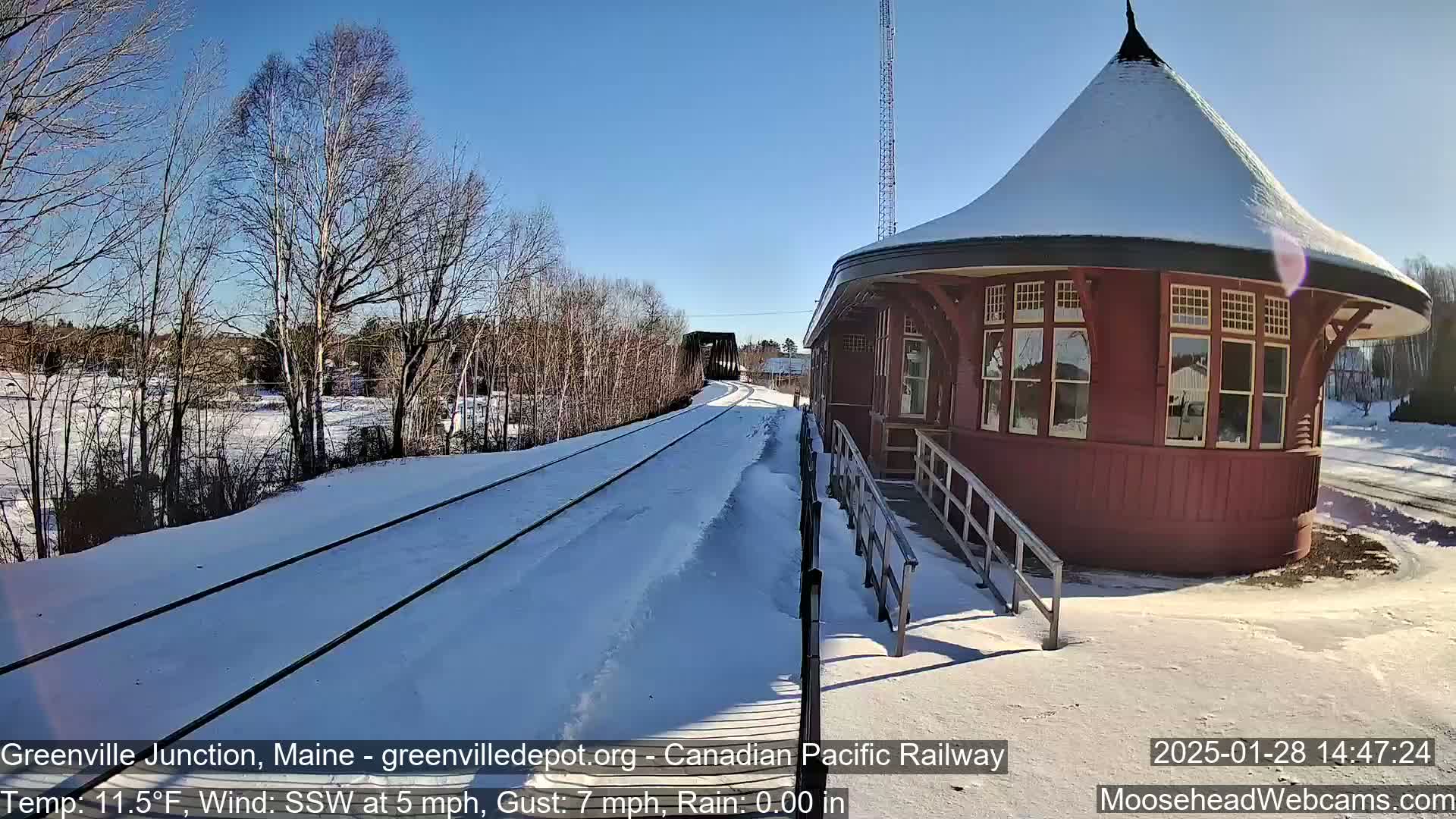 A snow-covered train station with a circular design sits next to train tracks on a sunny day with a light south-southwesterly wind.
