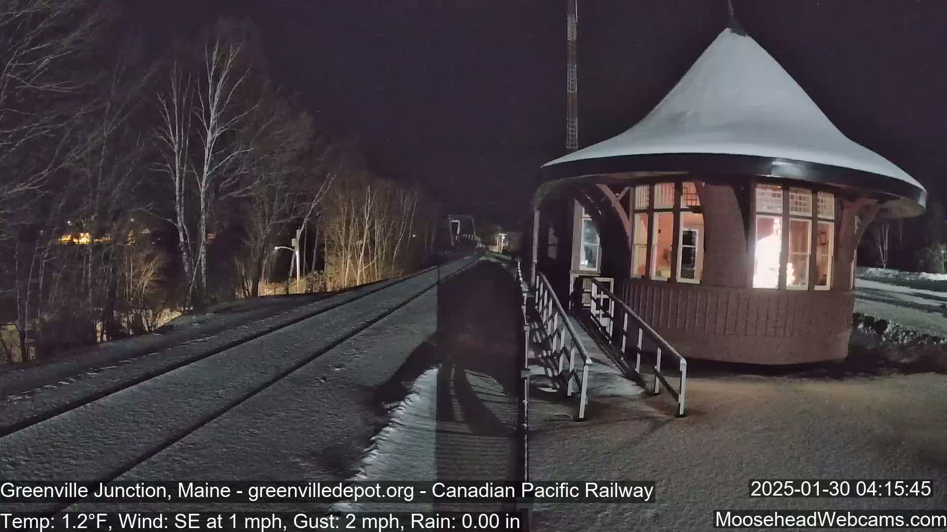 A snow-covered train platform and tracks are illuminated at night near a small, lit-up, snow-dusted building.