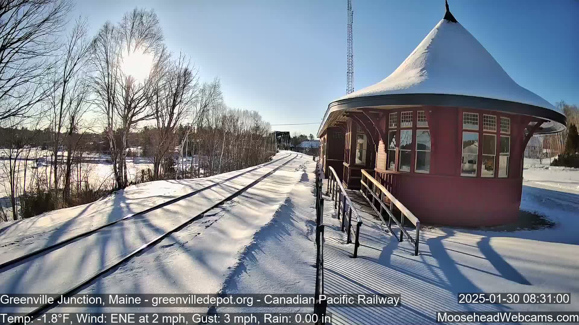 A snow-covered train platform with a red octagonal building and train tracks extends into the sunny winter landscape.