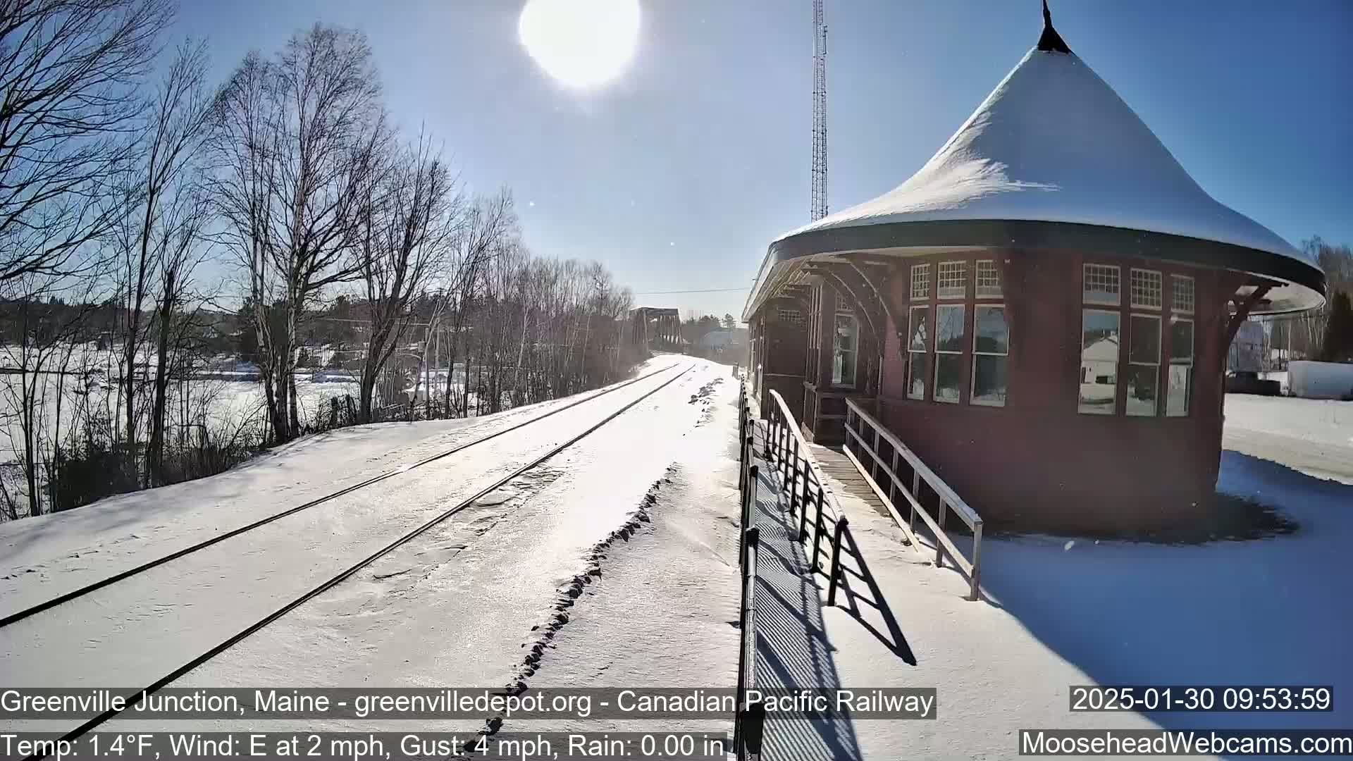 A snow-covered train track leads to a red, snow-dusted train station under a bright, sunny sky.