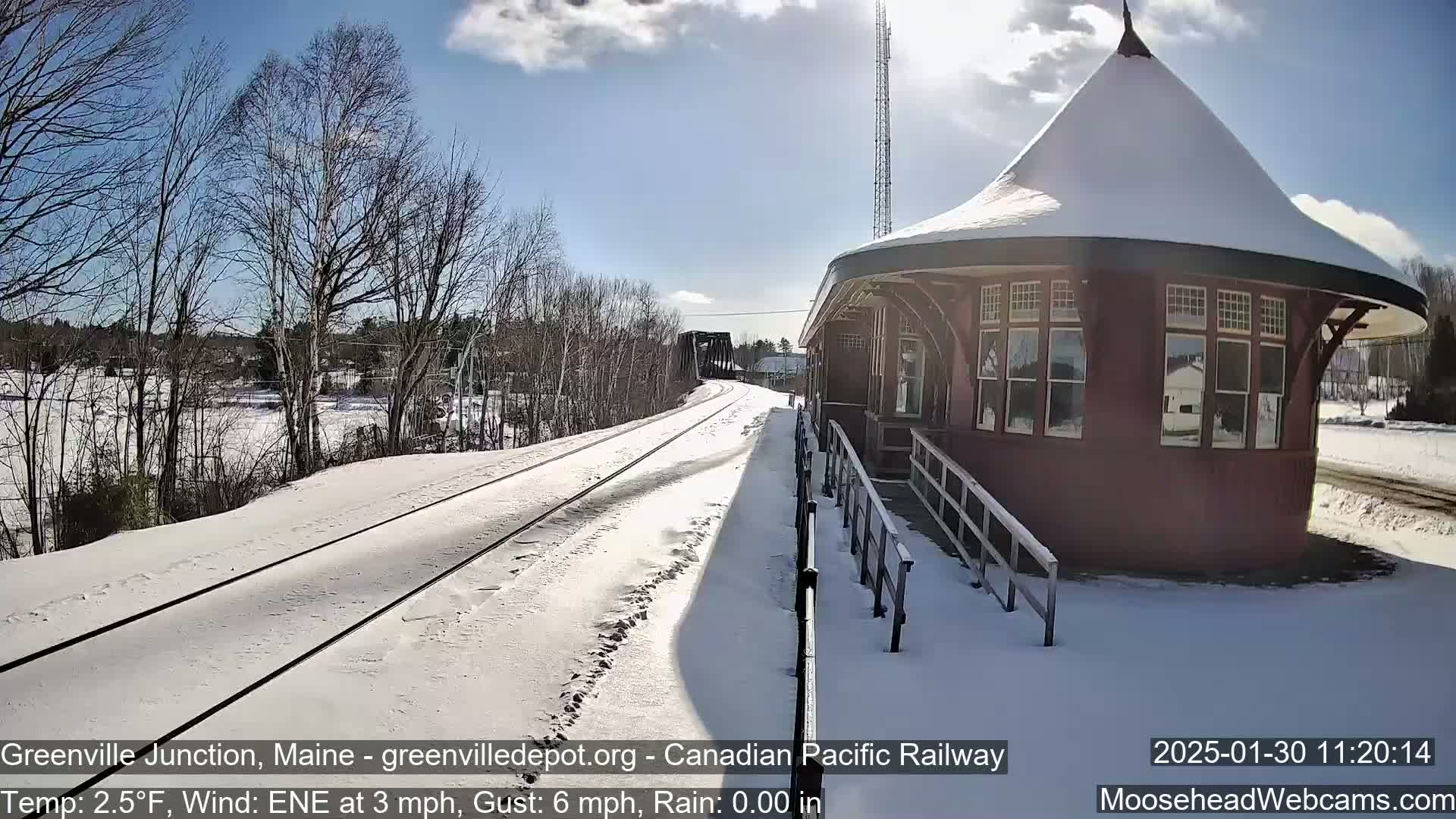 A snow-covered railway track leads to a snow-covered, octagonal train station under a partly sunny sky.