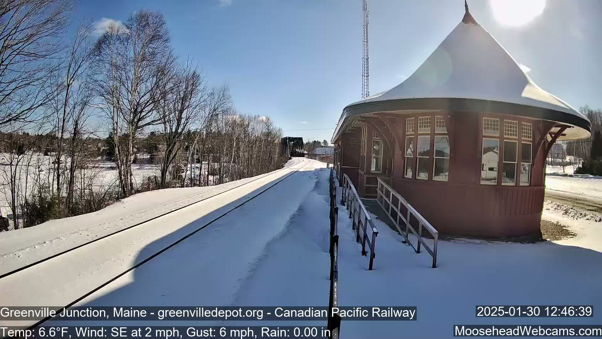 A snow-covered train platform and tracks extend toward a bridge under a sunny sky.