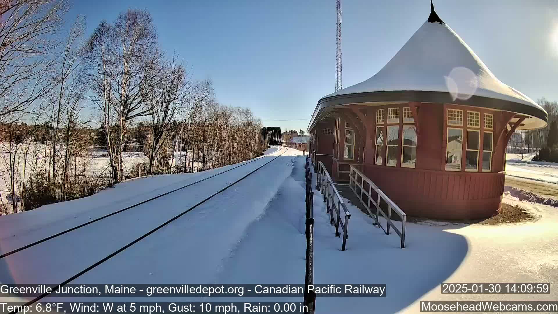 A snow-covered train platform with a red, round train station is visible on a sunny, cold day with a westerly wind.
