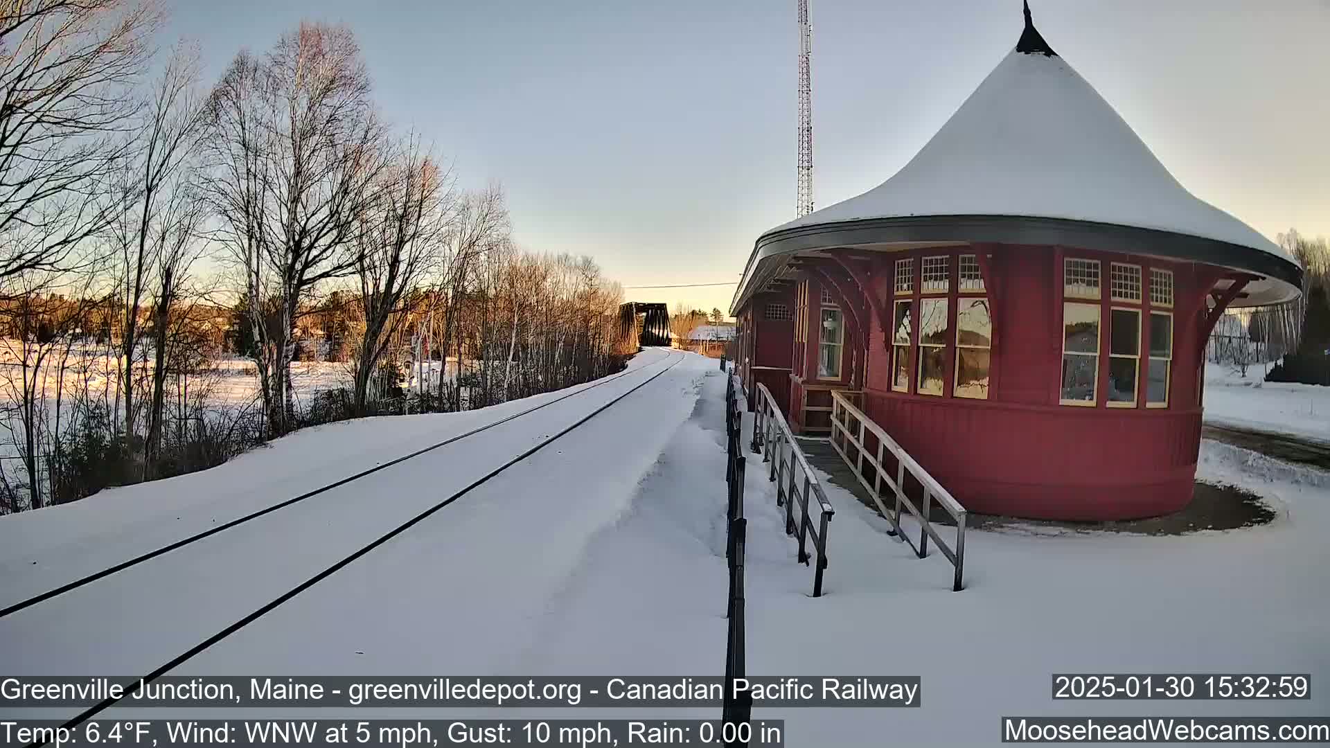 A snow-covered train track leads to a red, round train depot under a mostly clear sky.