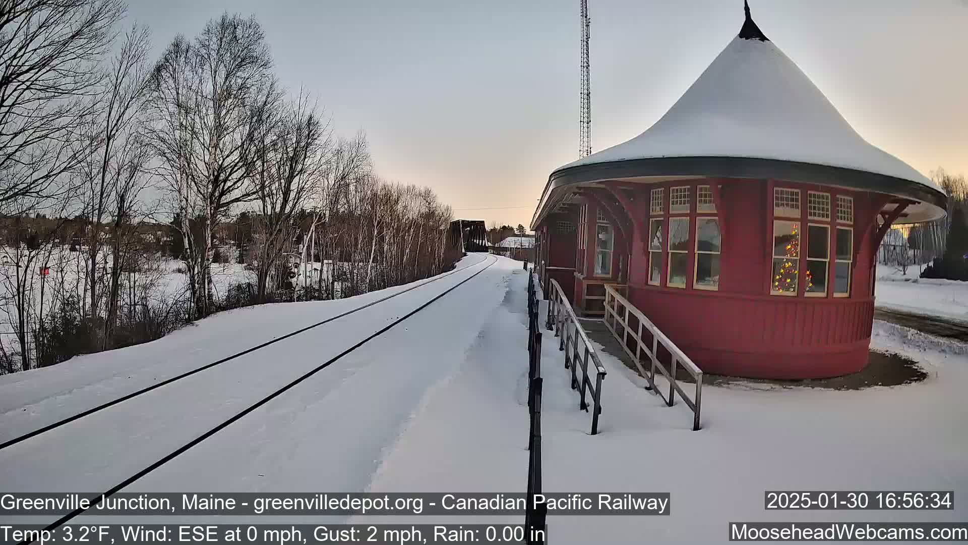 A snow-covered railway track leads to a red, round train station with a Christmas tree visible in its window, under a twilight sky.