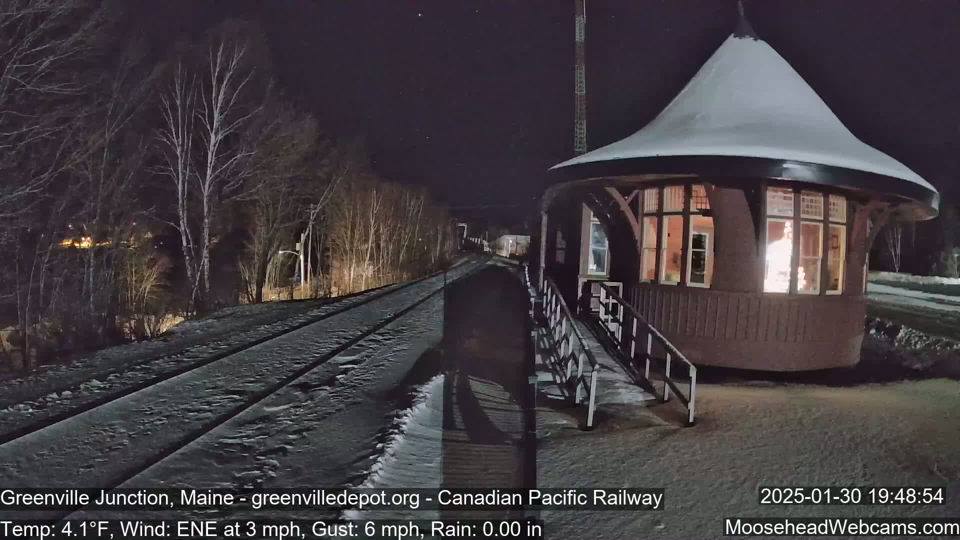 A snow-covered train track leads to a small, illuminated, snow-covered train station at night.