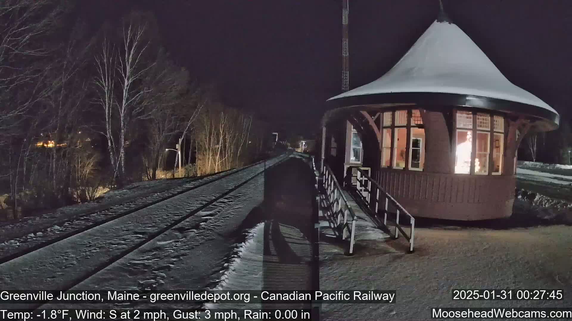 A snow-covered train track at night leads to a lit-up, snow-dusted, octagonal train station under a dark sky.
