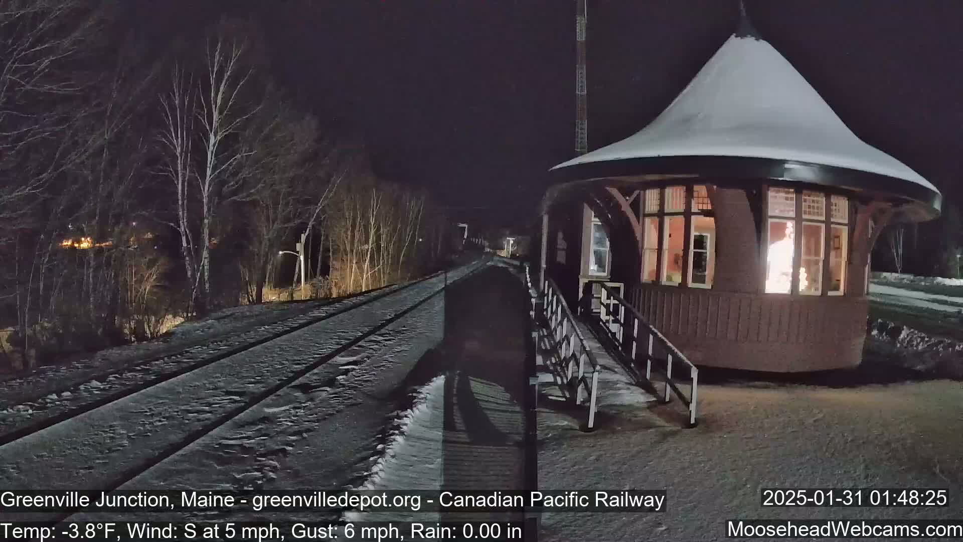 A snow-covered train station with a lit interior sits beside a snow-covered railway track at night.