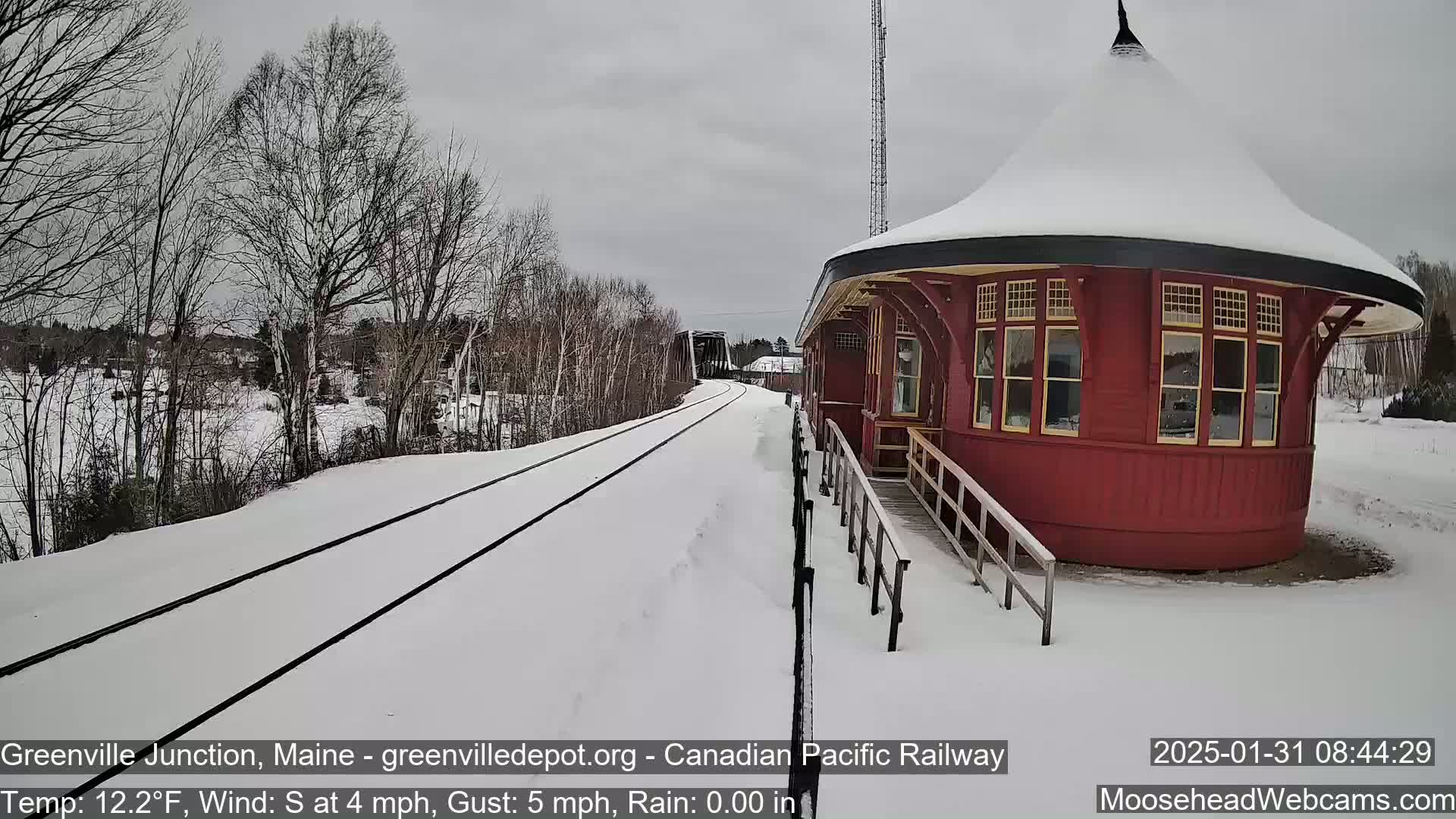 A snow-covered railway track leads to a round, red train station building under a gray sky.