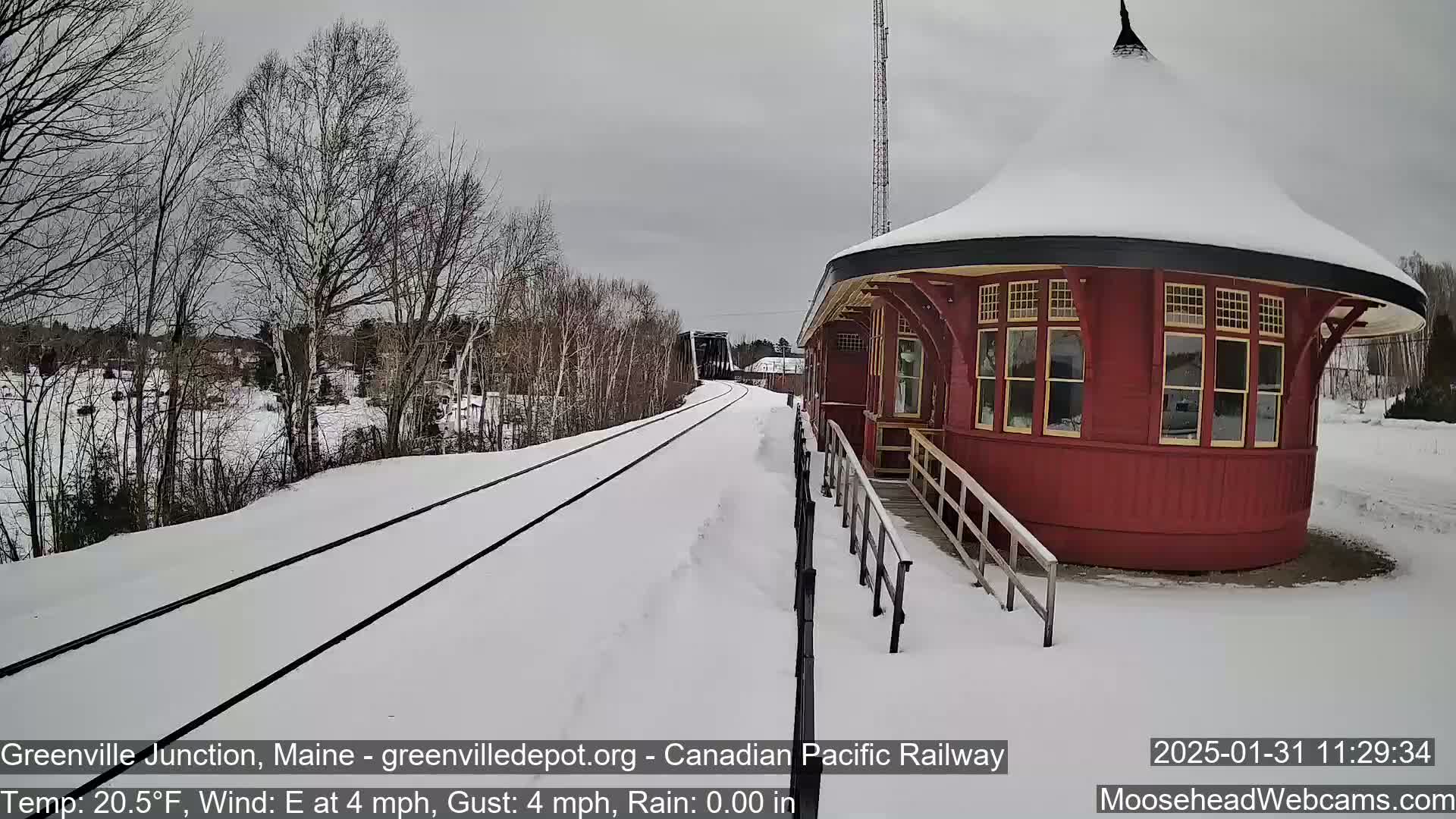 A snow-covered train track leads to a red, round train station with a snow-covered roof, under a cloudy sky.